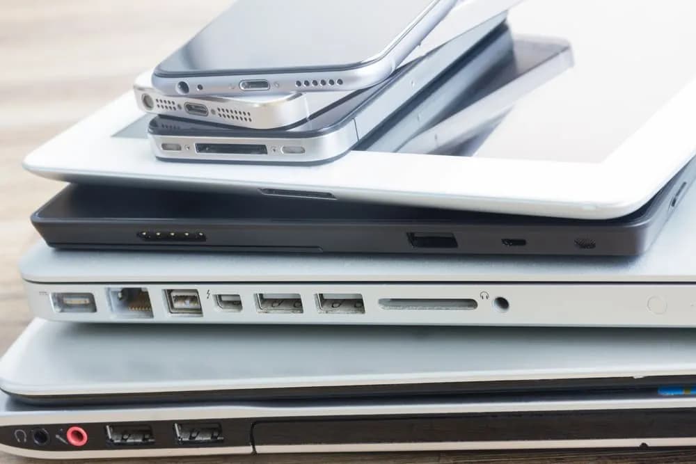 Stack of electronic devices including smartphones, a tablet, and several laptops piled on top of each other.