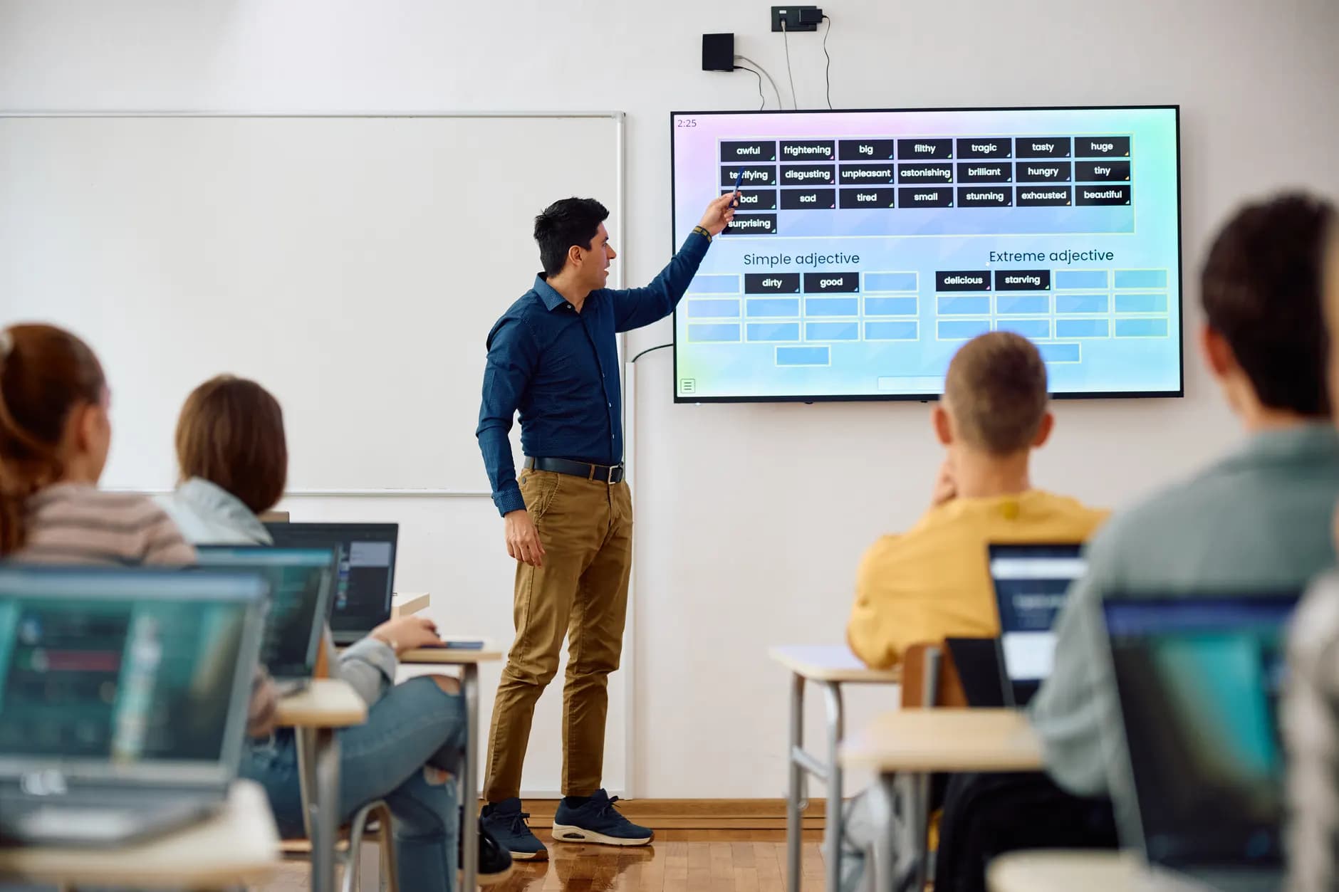 Teacher pointing to an interactive display while students work on laptops.